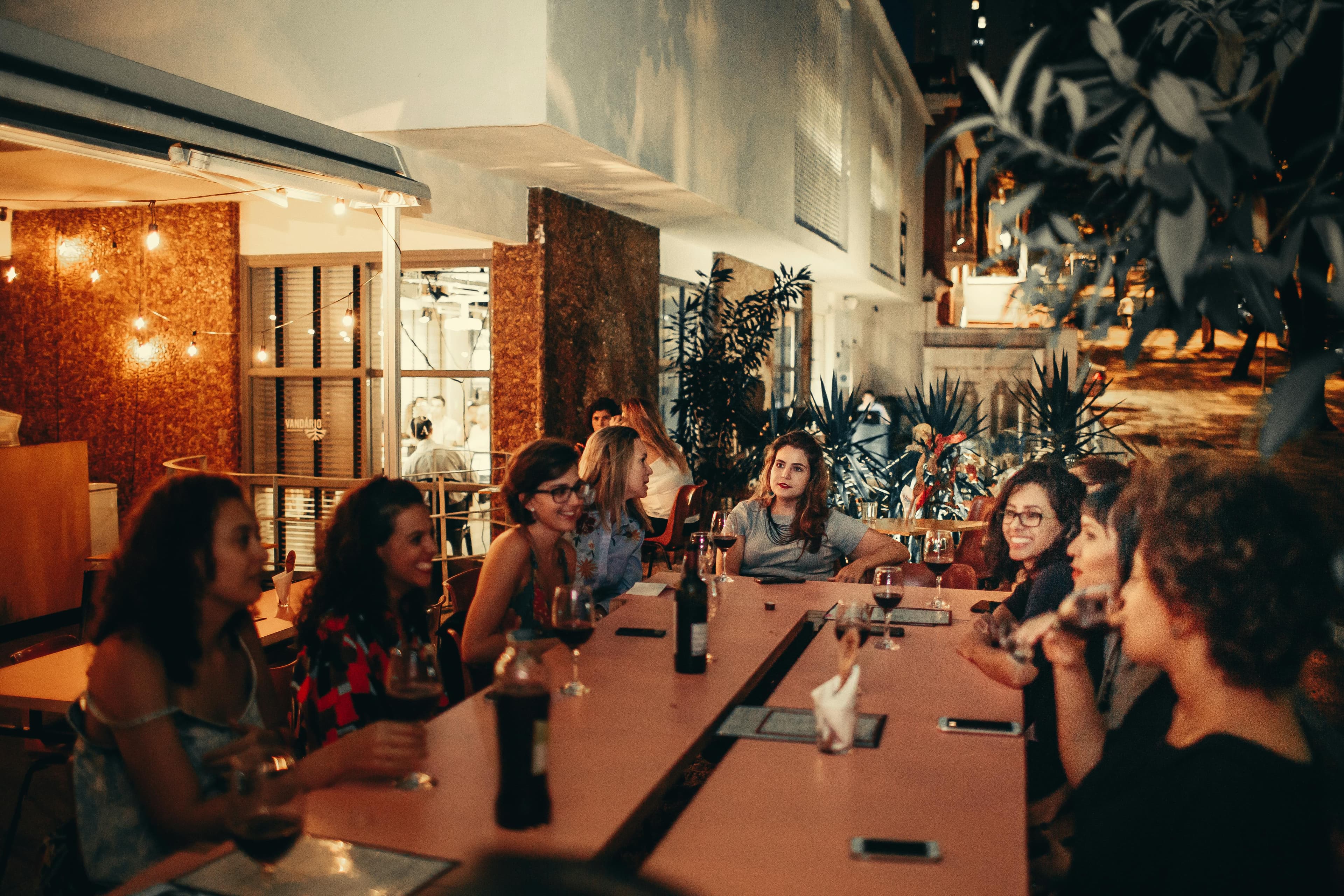 A long restaurant table at golden hour, two crews mid-conversation over wine